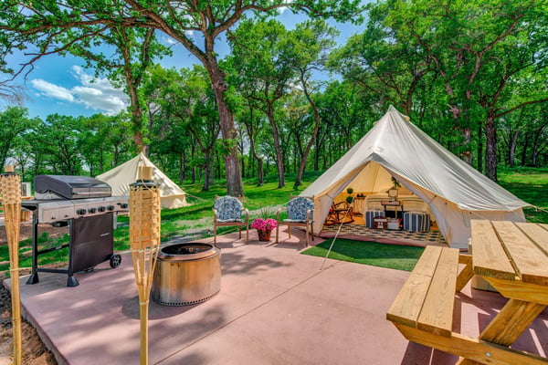 Glamping site with canvas bell tents, fire pit, grill, and wooden seating surrounded by tall trees
