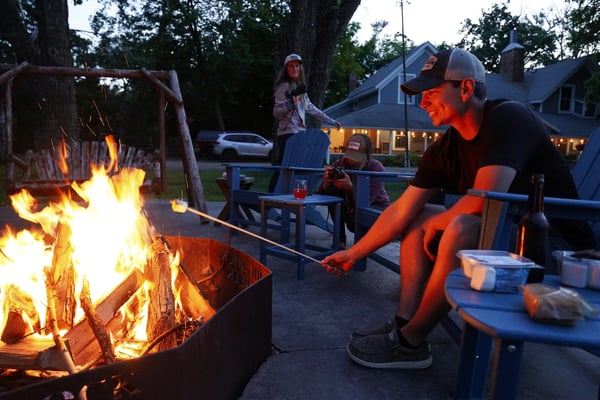 Family gathered around a backyard fire pit at dusk, roasting marshmallows with a house and trees in the background.